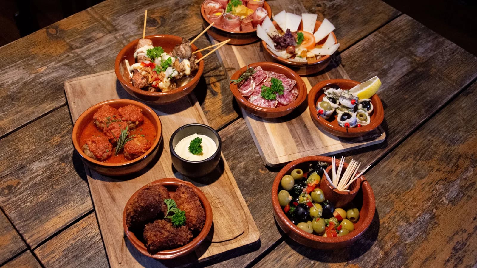 Tapas españolas para compartir en una mesa rústica en Tenerife: aceitunas, albóndigas, croquetas, embutido y queso en cazuelas de barro