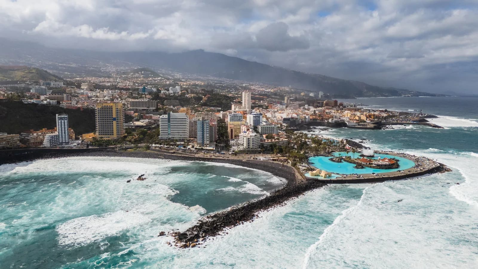 Vista aérea de Puerto de la Cruz, Tenerife: las piscinas turquesas del Lago Martiánez sobre un promontorio, la playa negra y la ciudad bajo las laderas del valle de La Orotava