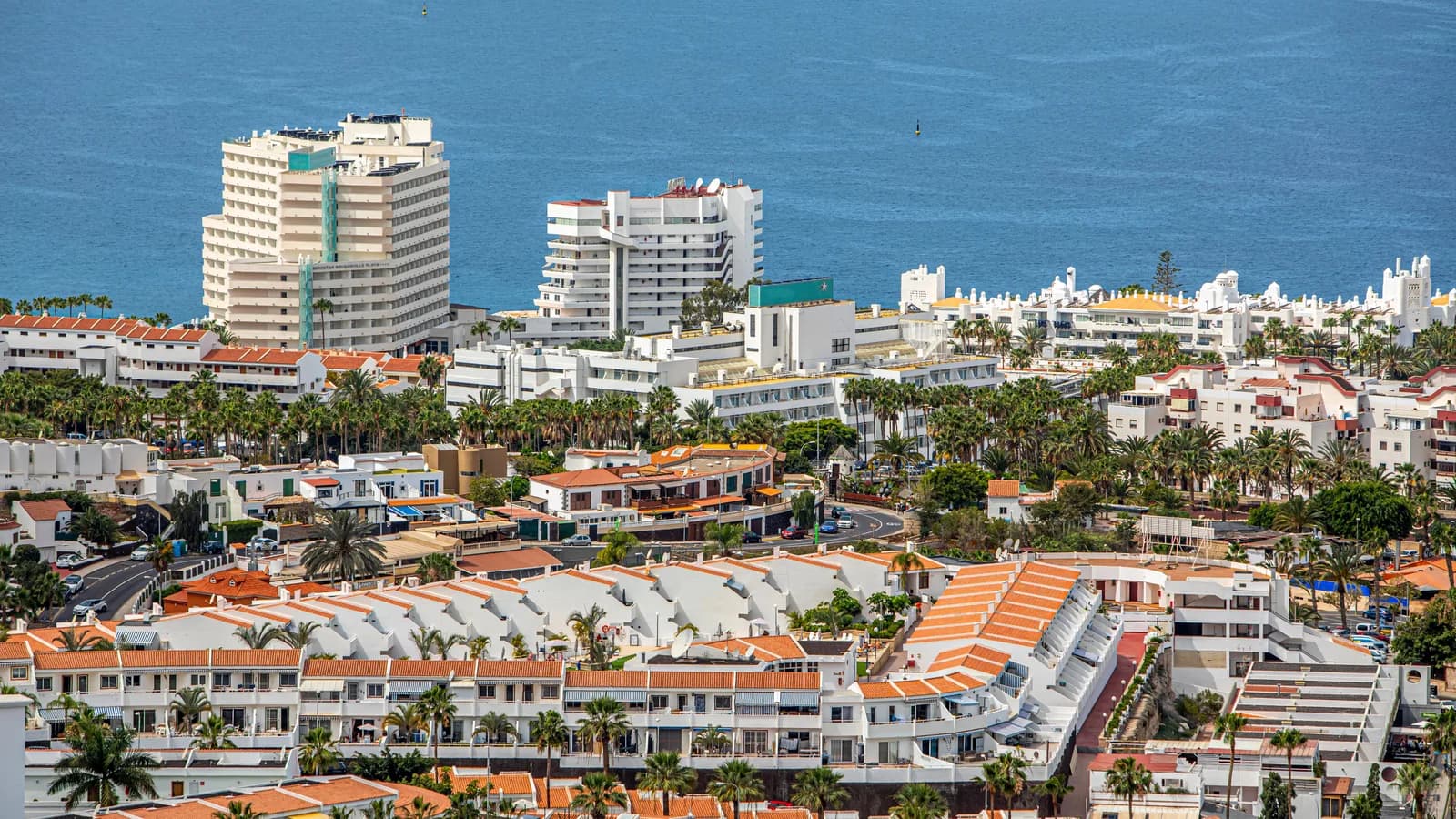 Vista aérea de la zona turística del sur de Tenerife: edificios blancos con tejados anaranjados, palmeras y el océano Atlántico al fondo